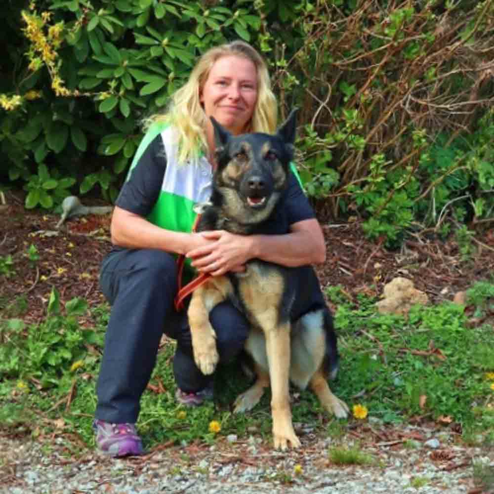 Hundetrainer steht mit einem sitzenden Deutschen Schäferhund auf einer Wiese im Freien