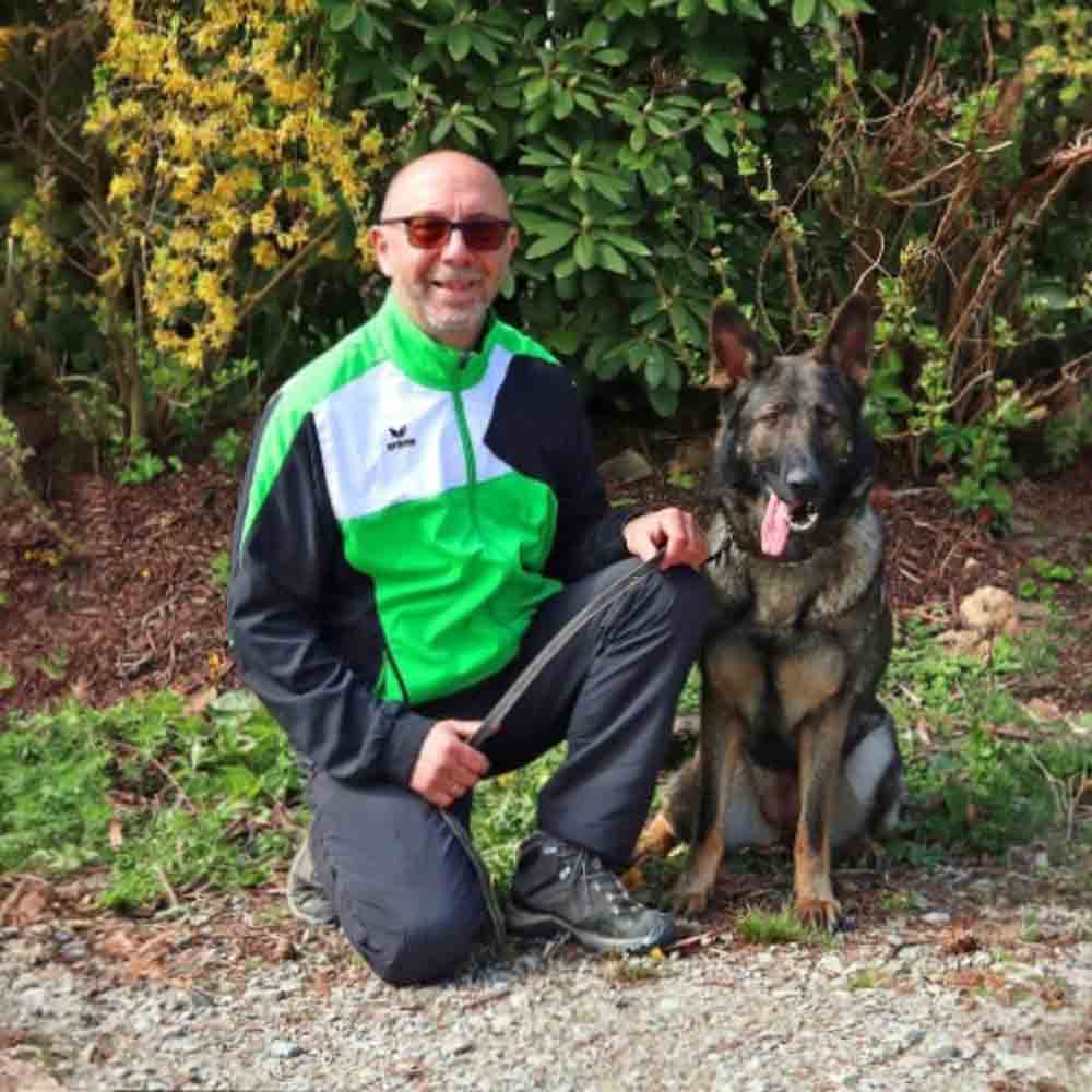 Hundetrainer steht mit einem sitzenden Deutschen Schäferhund auf einer Wiese im Freien