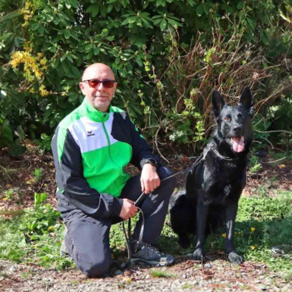 Hundetrainer steht mit einem sitzenden Deutschen Schäferhund auf einer Wiese im Freien