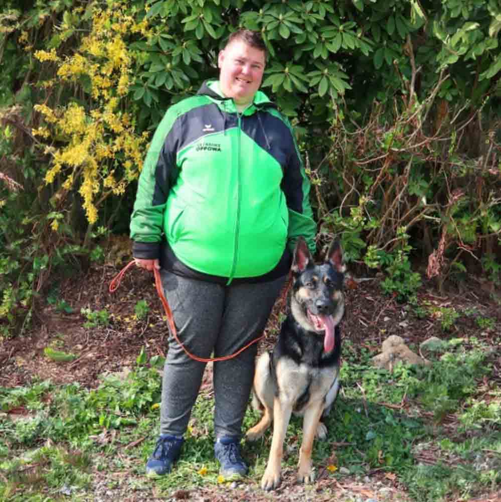 Hundetrainer steht mit einem sitzenden Deutschen Schäferhund auf einer Wiese im Freien